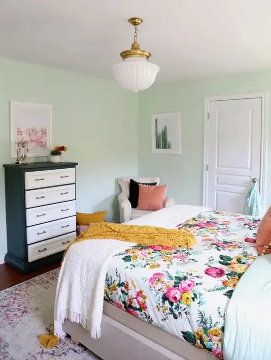 a mint-colored bedroom with a bed and floral bedding, a black and white dresser, a neutral chair and a chandelier