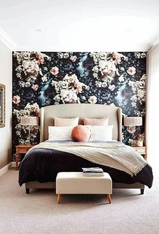 a modern refined bedroom with a black and blush floral accent wall, a neutral upholstered bed, contrasting bedding, an upholstered stool
