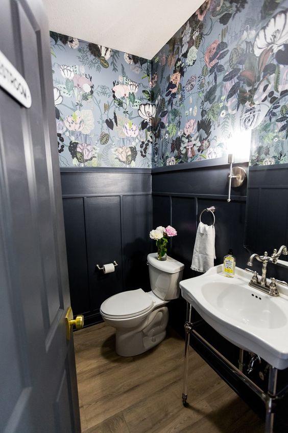 a moody powder room with grey floral wallpaper and black panling, a free-standing sink and a toilet, some blooms