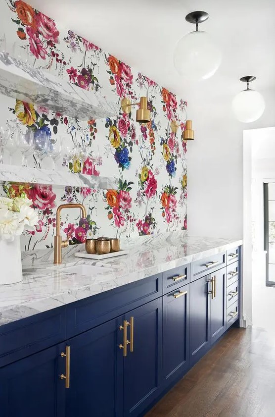 a navy and white kitchen with shaker cabinets, white marble countertops, a bright floral wallpaper wall and open shelves plus brass touches