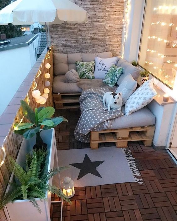 A small and cozy balcony with a pallet L shaped sofa, printed pillows, plants in pots, lights and candle lanterns