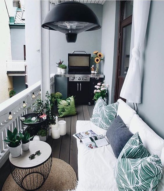 A welcoming balcony with a white L shaped sofa, printed pillows, a grill, potted greenery and blooms