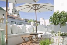 a white summer terrace with a built-in bench, white pillows, pottted blooms and greenery and umbrellas