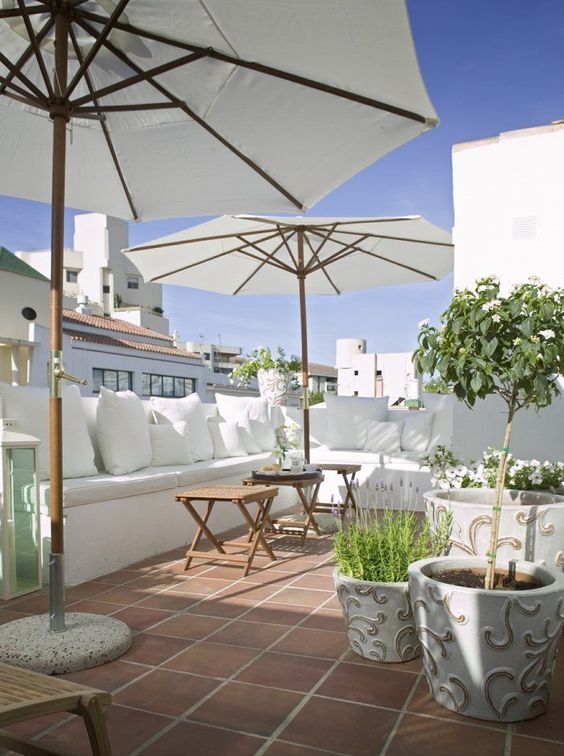A white summer terrace with a built in bench, white pillows, pottted blooms and greenery and umbrellas