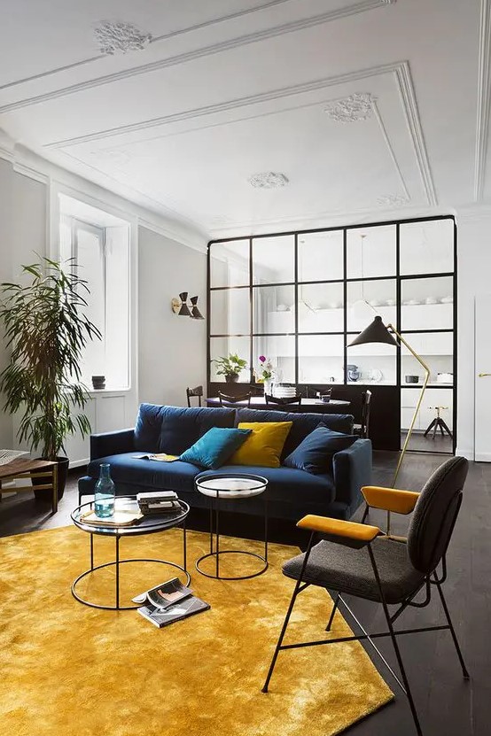 An elegant living room with a navy sofa, a black and yellow chair, a yellow hexagon shaped rug and pillows plus potted plants