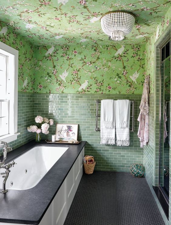 an eye-catchy green bathroom with floral wallpaper and green tiles, a bathtub with paneling, a bead chandelier and some blooms