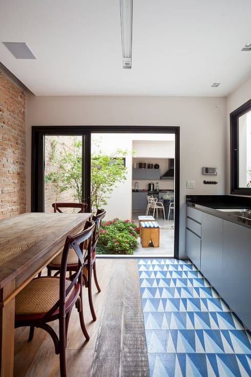 An indoor outdoor kitchen with sleek grey cabinets and black countertops, a blue and navy geo tile floor and a dining zone with wooden furniture
