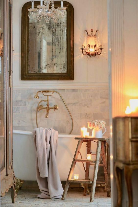 a Provence bathroom with marble tiles, a clawfoot bathtub, a vintage mirror, crystal lamps and candles and vintage fixtures