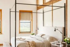 a Provence style bedroom with wooden beams, a series of windows for natural light, a metal canopy bed with grey bedding, round nightstands and a wooden bench