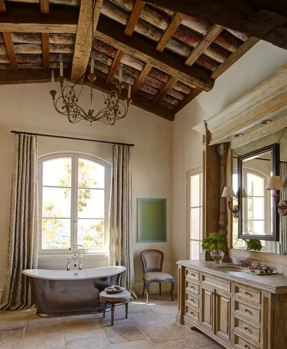 a beautiful Provence bathroom with a wooden ceiling and wooden beams, a large wooden vanity, limewashed walls, a shiny metal bathtub and vintage furniture