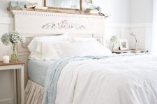 a chic Provencal bedroom with white paneling, a bed with a refined headboard, blue and white bedding, books and a mirror, a crystal chandelier