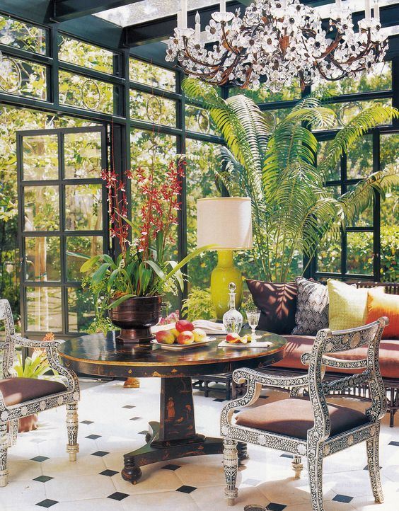 a colorful vintage sunroom with a dark stained table, refined chairs, a daybed with vibrant pillows, potted greenery and a gorgeous floral chandelier