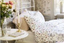 a dreamy Provence bedroom with floral wallpaper, a white metal bed with floral bedding, a white table, a floral bench, artwork and blooms