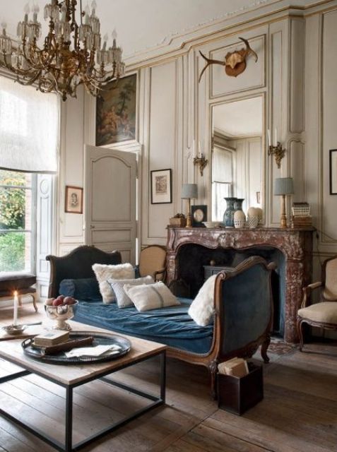 a jaw-dropping living room with neutral walls and paneling, a fireplace clad with marble, a blue daybed, a chic chandelier and artworks