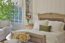 a refined French chic bedroom with shiplap, a refined bed with cane touches, lovely seating furniture, a crystal chandelier and some greenery