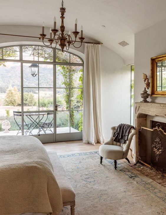 a refined French country chic bedroom with a fireplace and a cover, a bed with neutral bedding and a bench, a chandelier and a mirror in a gilded frame