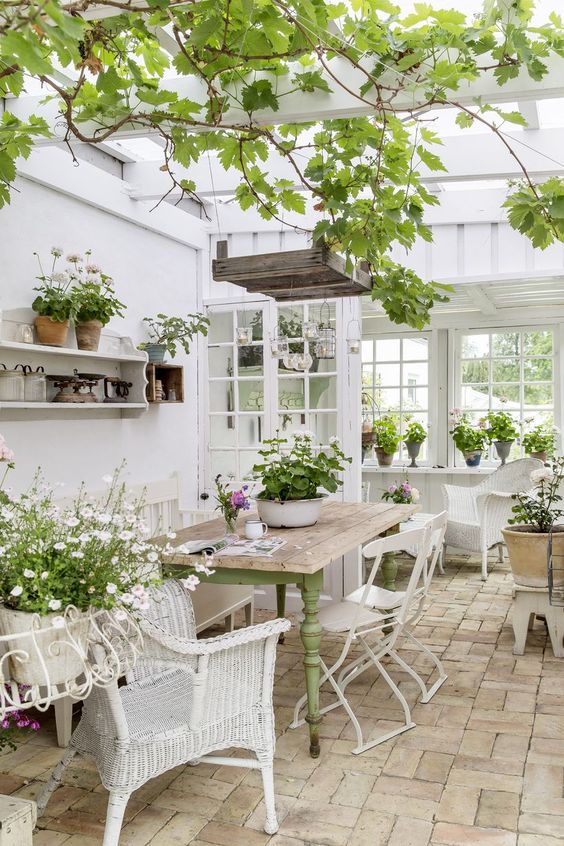 A refined vintage sunroom with white furniture   wicker and metal, a green table, potted greenery and blooms and hanging lamps