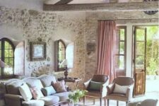 a rustic Provence living room with stone walls, wooden beams, refined vintage furniture, pink curtains and a pink ottoman as a table