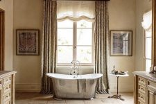 a soft buttermilk bathroom with textural stone floors and a wooden ceiling, a shiny metal bathtub, a beautiful metal chandelier and wooden vanity