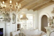 a sophisticated Provence living room with a fireplace, a crystal chandelier, a heavy table, neutral and refined furniture and wooden beams