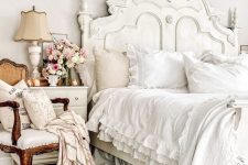a sophisticated neutral Provence bedroom with a refined bed and bedding, a dresser, a chic creamy chair, a lovely lamp and some blooms
