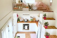 a tiny home with a loft bedroom, with some stands and storage units, a low bed with pastel bedding and some potted plants on the stairs