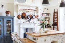 a vintage kitchen with white shabby chic cabinets, butcherblock countertops, black pendant lamps, a blue buffet and a vintage hearth