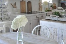 a white vintage kitchen with a shabby chic kitchen island, a wooden hood, pendant lamps and a crystal chandelier, a shabby chic dining set