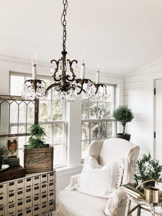 a white vintage sunroom with neutral furniture, a vintage storage unit, a crystal chandelider and potted greenery