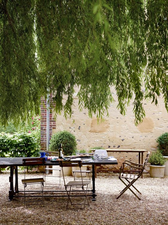 A Provence inspired outdoor space with a stained table and metal and wood chairs, greenery and trees over the space