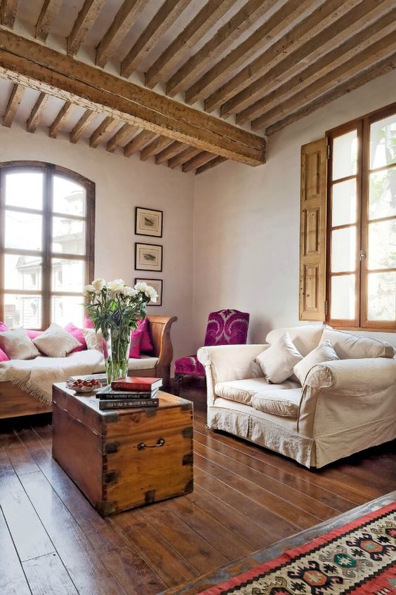 a Provence living room with wooden beams and shutters, neutral refined furniture, a chest in the center and a gallery wall