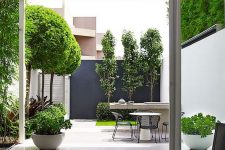 a catchy minimalist outdoor space with growing trees, grass and greenery, a stone table and metal chairs and a black accent wall