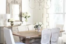 a charming French country dining room with a neutral sideboard, a stained vintage table, white chairs, a lovely chandelier and a mirror in a wooden frame
