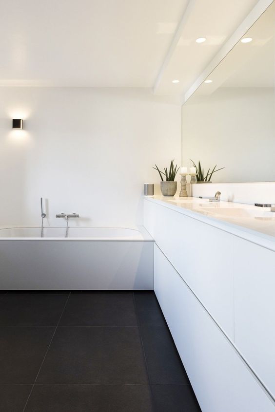 A contrasting black and white bathroom with a black tiled floor, a white long vanity, a bathtub clad in white and some built in lights