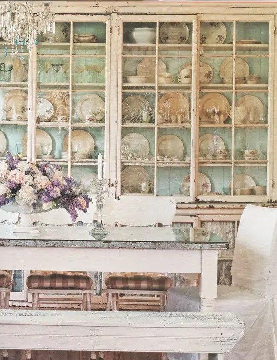 a gorgeous shabby chic dining room with a large shelving unit that takes a whole wall, shabby chic vintage furniture and a crystal chandelier