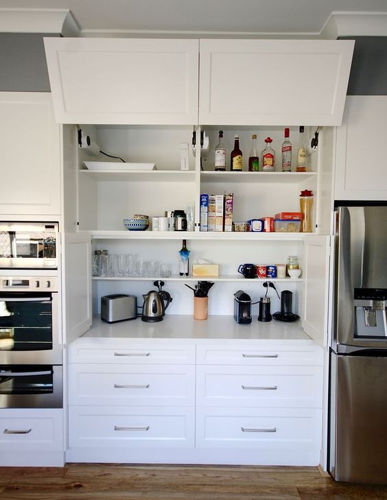 A large cabinet with open shelves and a garage style door is a home bar hidden with a kettle, a toaster and some syrups