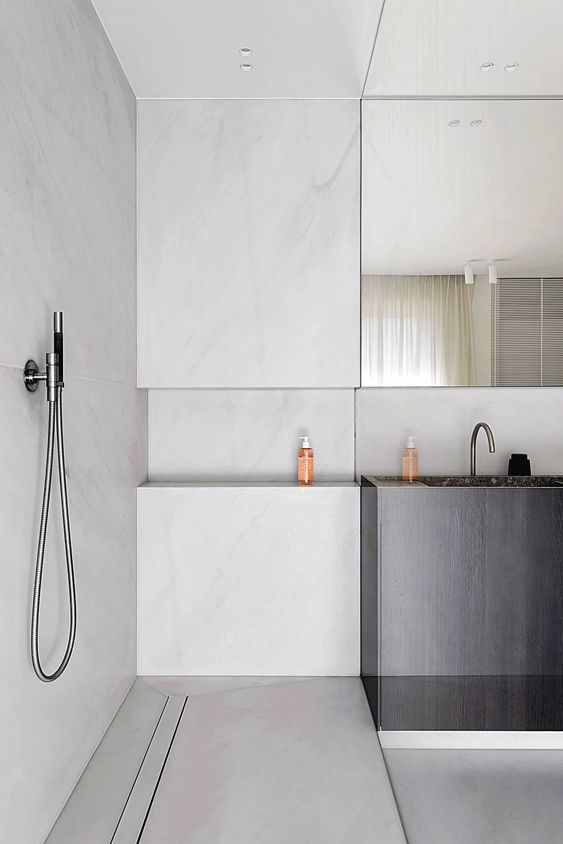 A minimalist bathroom clad with white marble, a dark vanity with a built in sink and a statement mirror coming up the ceiling