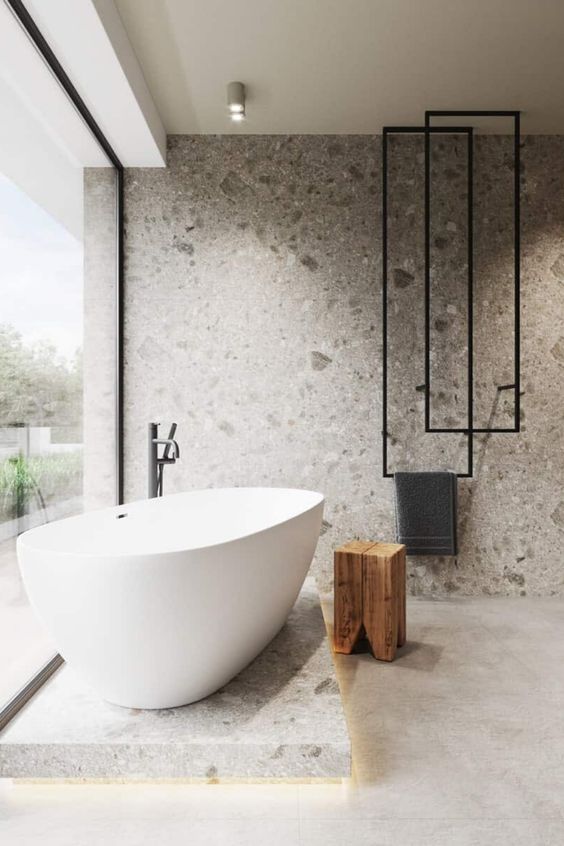 a minimalist bathroom with a glass wall, a bathtub on a stone platform, a stone wall and cool towel hangers
