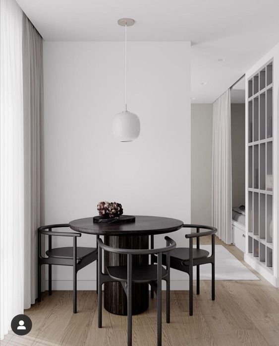 a minimalist black and white dining nook with white walls, a round black table and matching black chairs and a white pendant lamp