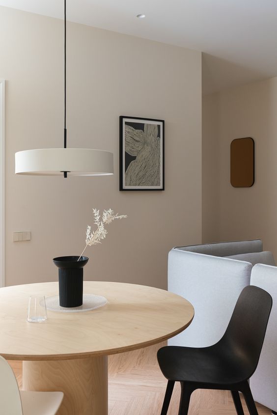 A minimalist dining space with a round light stained table, a black and neutral chair, a neutral pendant lamp and a black vase