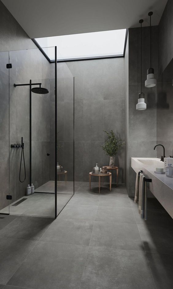 A minimalist grey stone like tiles, a shower space clad with glass, a floating vanity with a sink and pendant lamps plus a skylight
