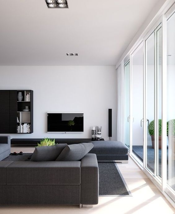 A minimalist living room with grey furniture, a TV, a wall mounted storage unit and a glazed wall for much natural light