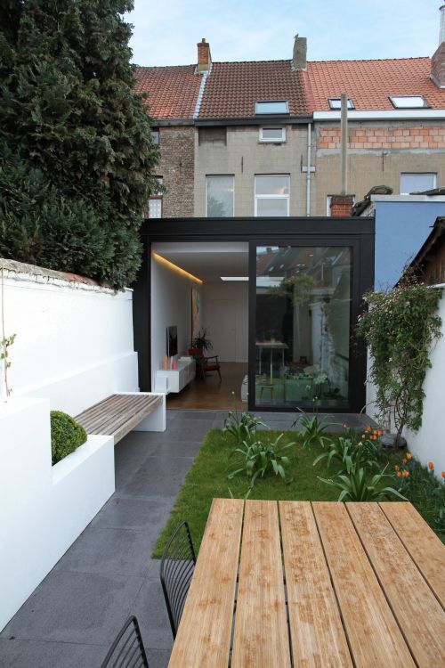 A minimalist terrace clad with tiles, built in bnches and potted greenery and blooms, a planked woodne table and metal chairs