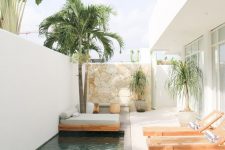 a minimalist terrace wiht a stone clad wall, wooden loungers, a daybed with a grey mattress with pillows, trees and a long pool