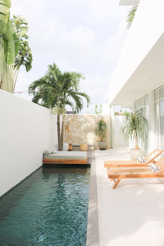 a minimalist terrace wiht a stone clad wall, wooden loungers, a daybed with a grey mattress with pillows, trees and a long pool