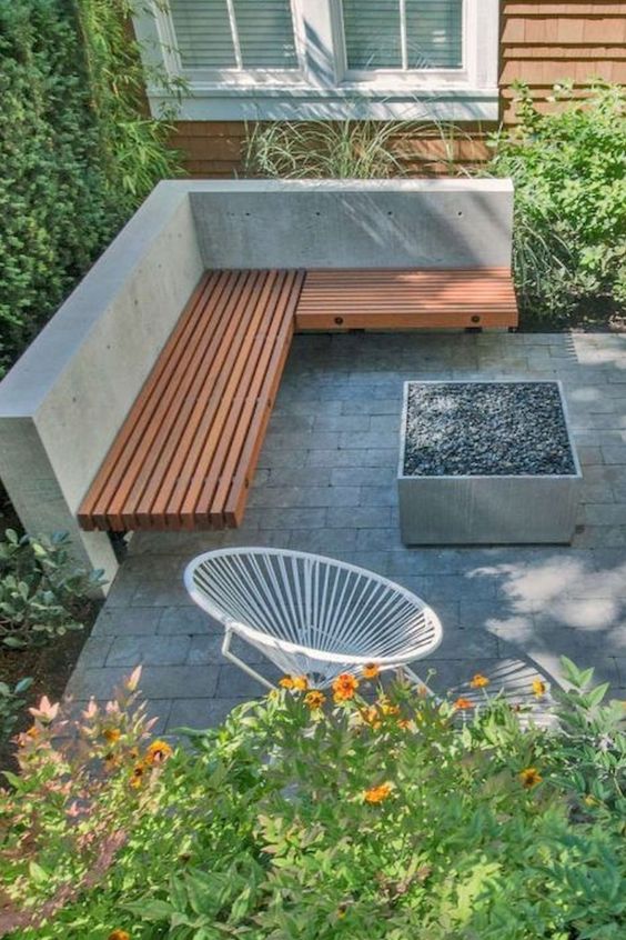 A minimalist terrace with concrete tiles, a built in corner bench, a fire pit, a round chair and lots of greenery and blooms around