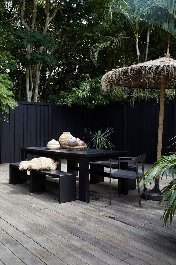 a minimalist tropical terrace with a reclaimed wooden deck, laconic black dining set and a tall umbrella plus vases on the table