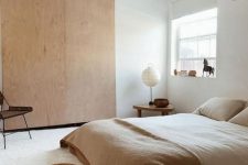 a natural and minimalist bedroom with a plywood storage unit, a bed with neutral bedding, wooden stools and a chair