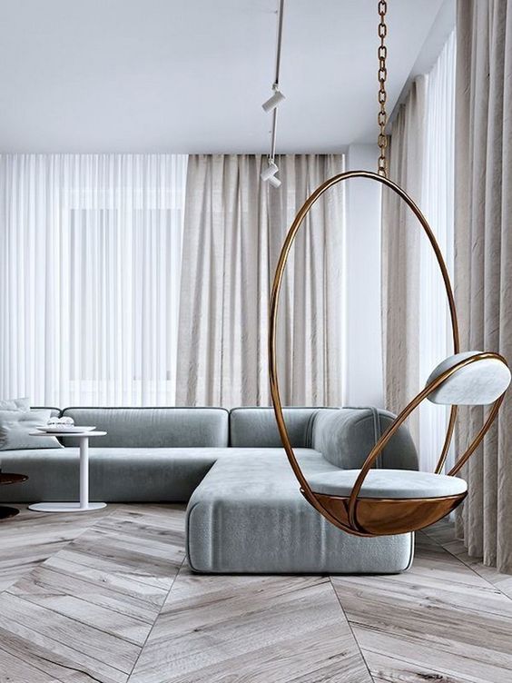 A neutral minimalist living room with off white curtains, a grey sectional sofa, a hanging copper chair and a laconic coffee table
