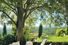 a refined French country terrace with black forged seating furniture, white upholstery and lots of greenery and a tree around